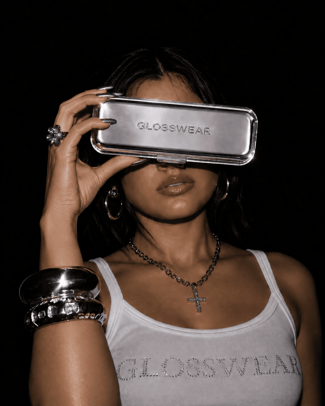 A woman holds a silver Glosswear case up to her eyes like a viewfinder, wearing a rhinestone Glosswear tank top.