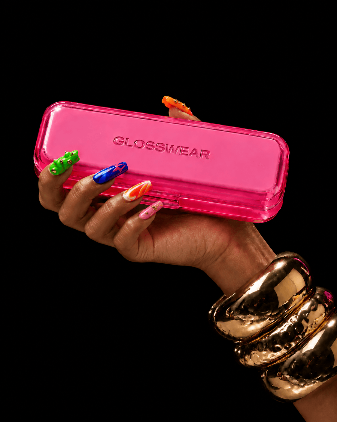 A hand with colourful press-on nails and gold bangles holds a pink translucent Glosswear case against a black background.