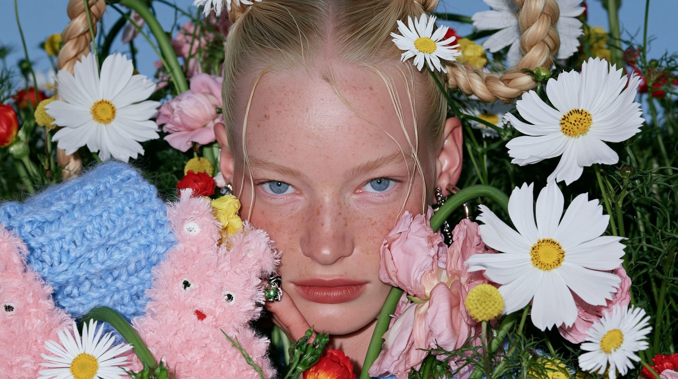 A close-up of a woman’s face surrounded by daisies as large as her head, a pink toy rabbit at her cheek.