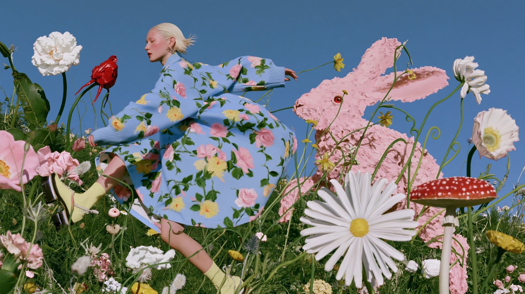A woman in a blue rose-print dress runs through a meadow past an oversized pink rabbit sculpture, a giant red rose and a toadstool beside her.