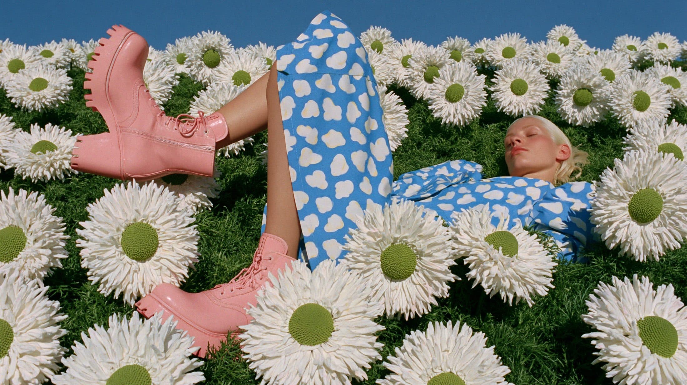 A woman lies asleep in a field of daisies grown to the height of her body, pink boots crossed in the air.