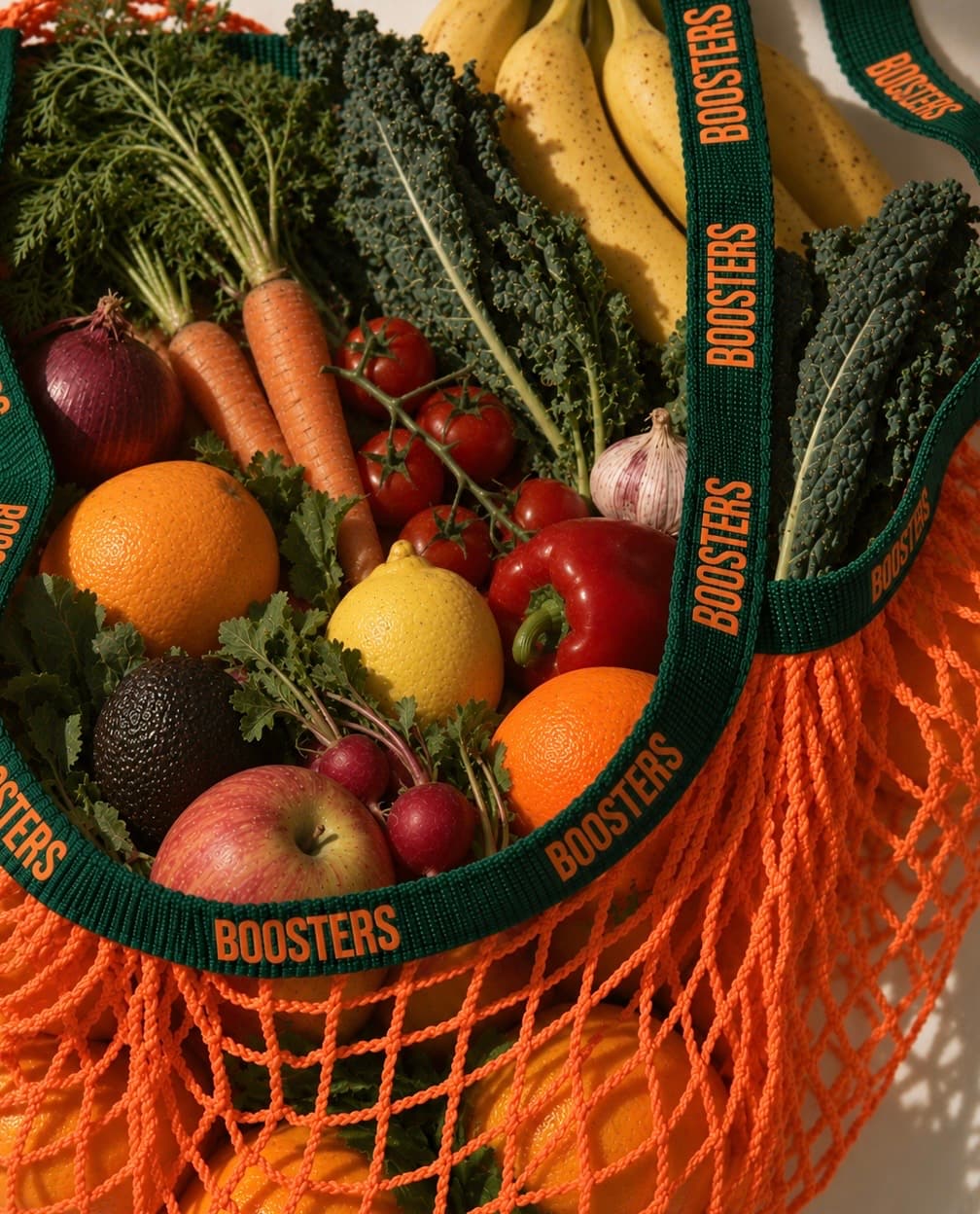 An orange string bag printed with the Boosters wordmark, packed with kale, tomatoes, citrus, carrots and bananas.