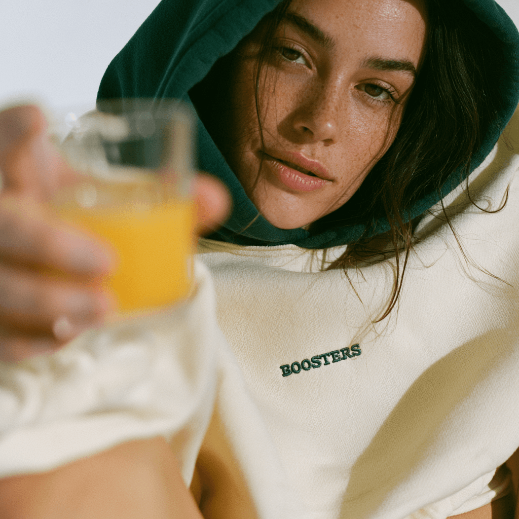 A woman in a cream and dark green Boosters Club hoodie holds out a glass of fresh orange juice.
