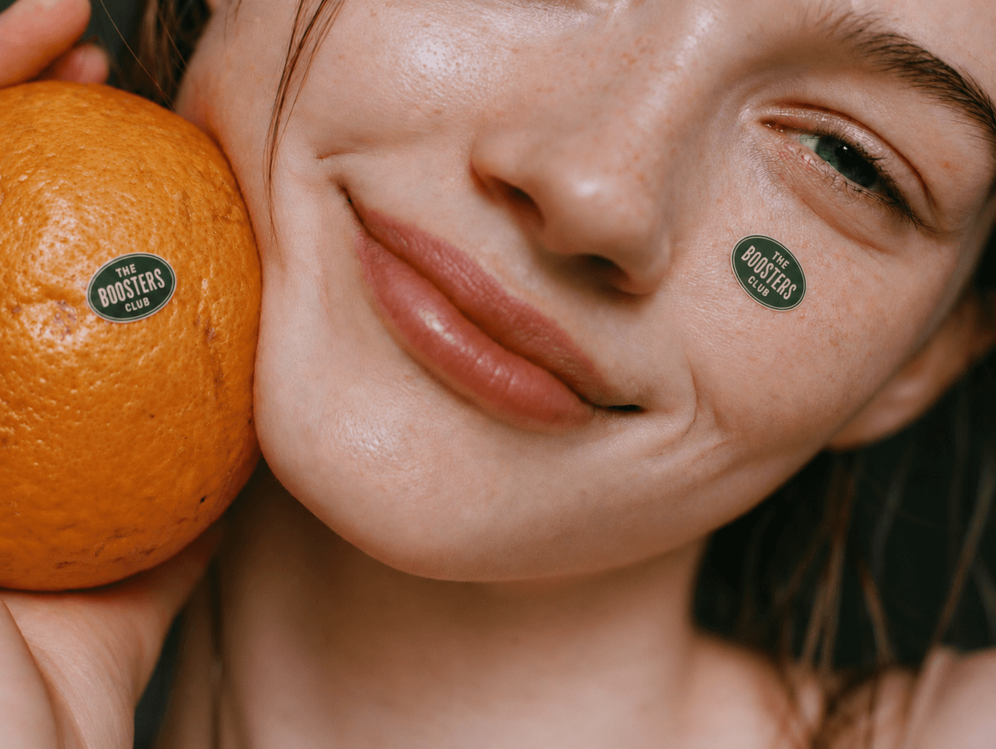 A woman holds an orange to her cheek, the small oval Boosters Club logo on both the fruit and her skin.