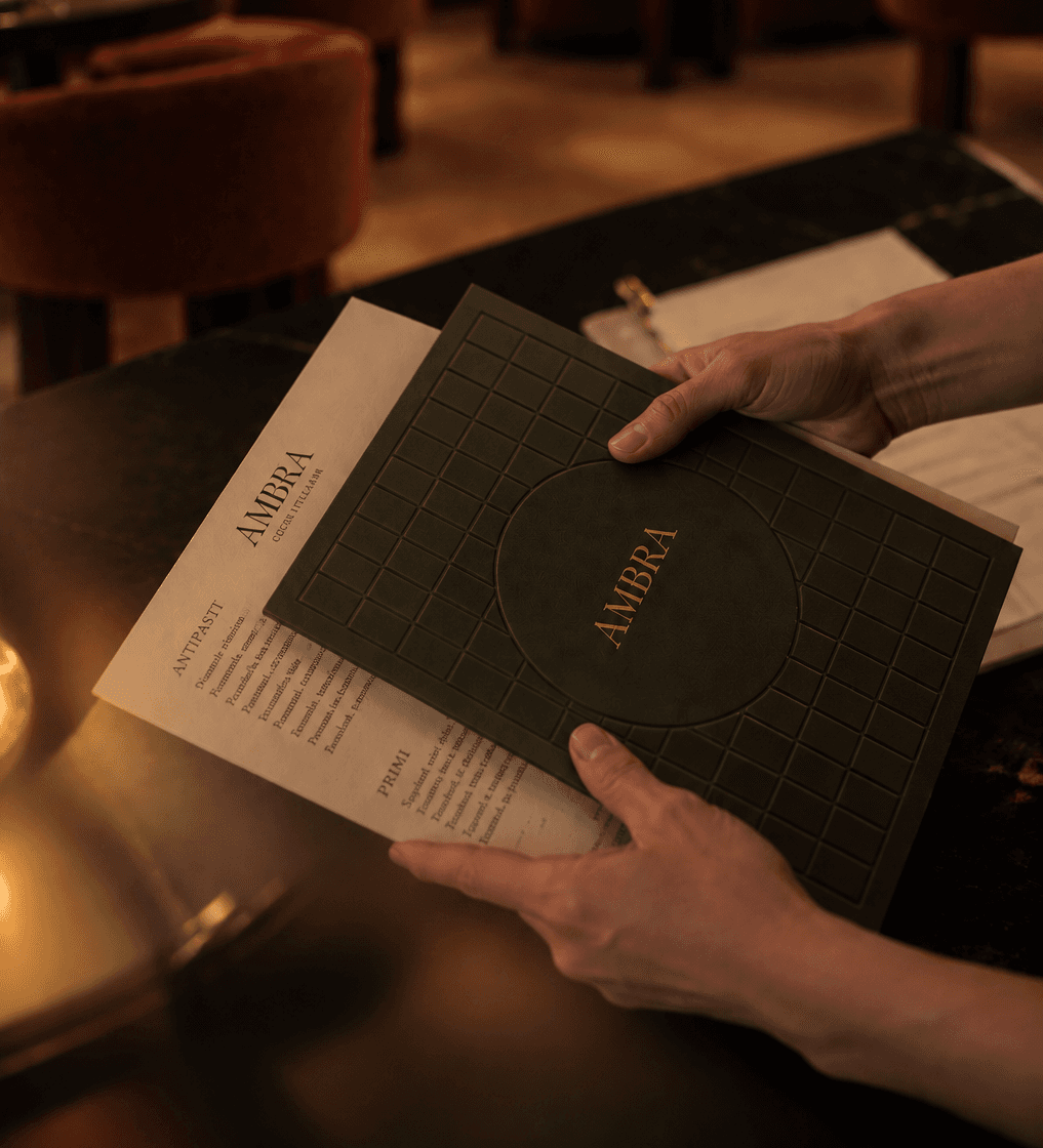 A waiter holding the Ambra menu open at the table, the debossed cover against the printed page.
