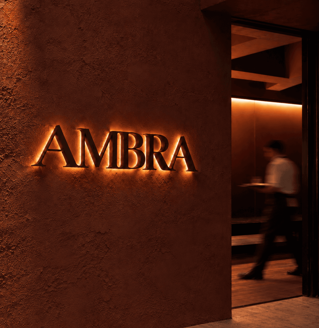 A lit amber sign outside Ambra reading AMBRA, Cucina Italiana, mounted on pale stone.