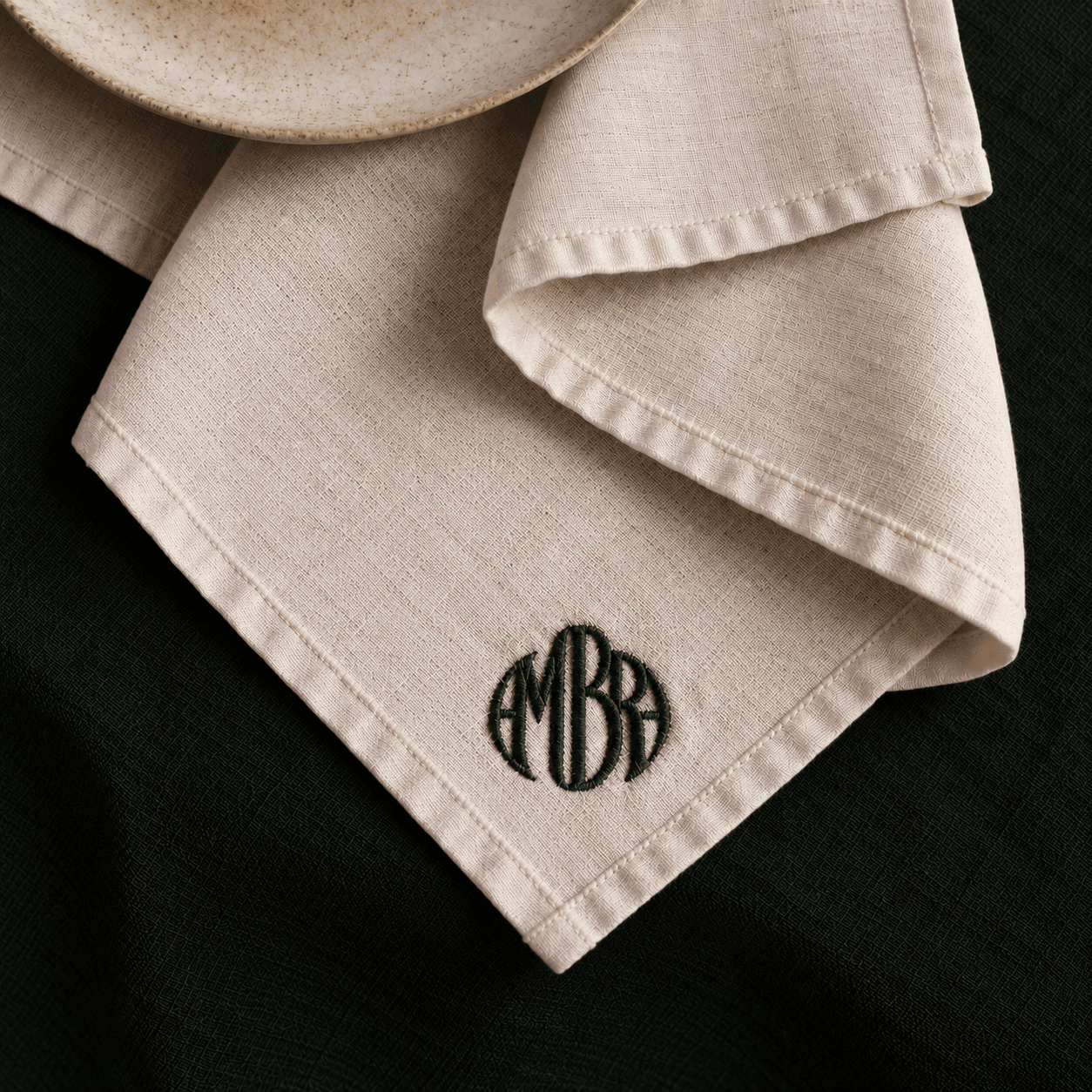 A linen napkin embroidered with the ambra wordmark, one corner of the amber circle motif printed across it, draped over a table edge.